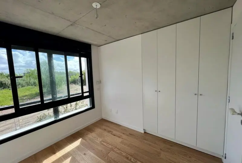 Puerta de acceso con amplio ventanal con vistas al exterior, habitación luminosa con armario empotrado y suelo de madera natural en proyecto de vivienda moderna en construcción.