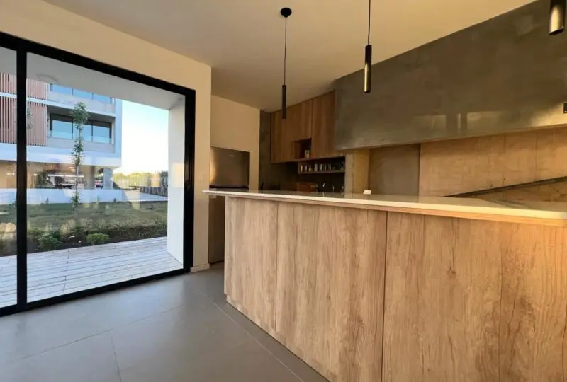 Moderno espacio interior de apartamento en venta, con cocina de diseño minimalista, encimeras de madera natural y grandes ventanales con vista a área verde exterior, ideal para inversión inmobiliaria de lujo.