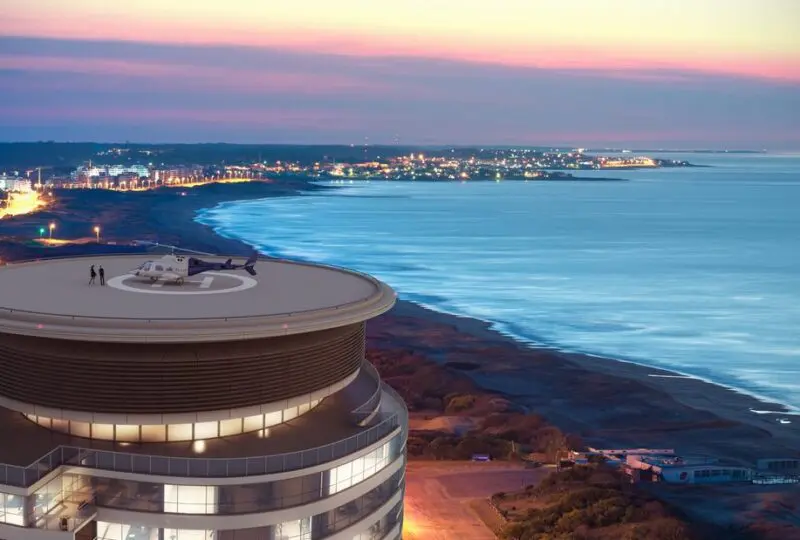 Vistas aéreas de un edificio moderno con helipuerto en la azotea, junto a la costa luminosa durante el atardecer en la ciudad.