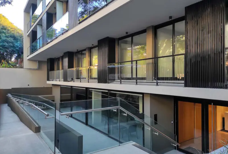 Moderno edificio residencial en altura con ventanas de piso a techo y balcones con barandillas de cristal, diseño arquitectónico innovador y elegante en zona urbana.